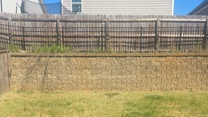 Un jardin avec un mur en briques, une clôture en bois et une maison en arrière-plan.