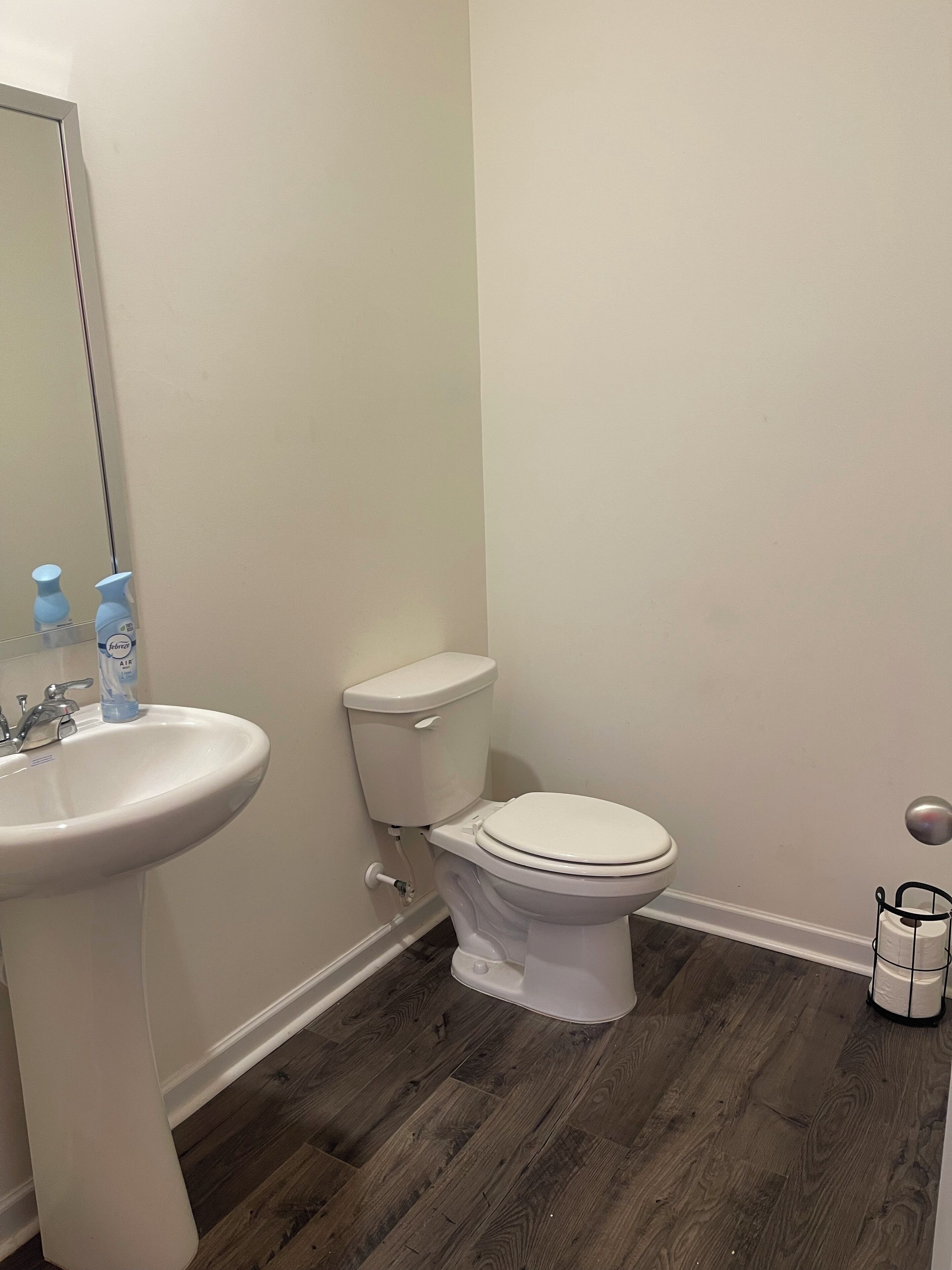 A bathroom with a white sink, toilet, and mirror. There are two bottles of air freshener on the sink. The floor is wooden.