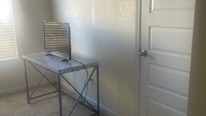Une pièce dont le sol est recouvert d’une moquette, avec une table à structure métallique et un dessus orné d’un motif, une fenêtre avec des stores et une porte blanche.
