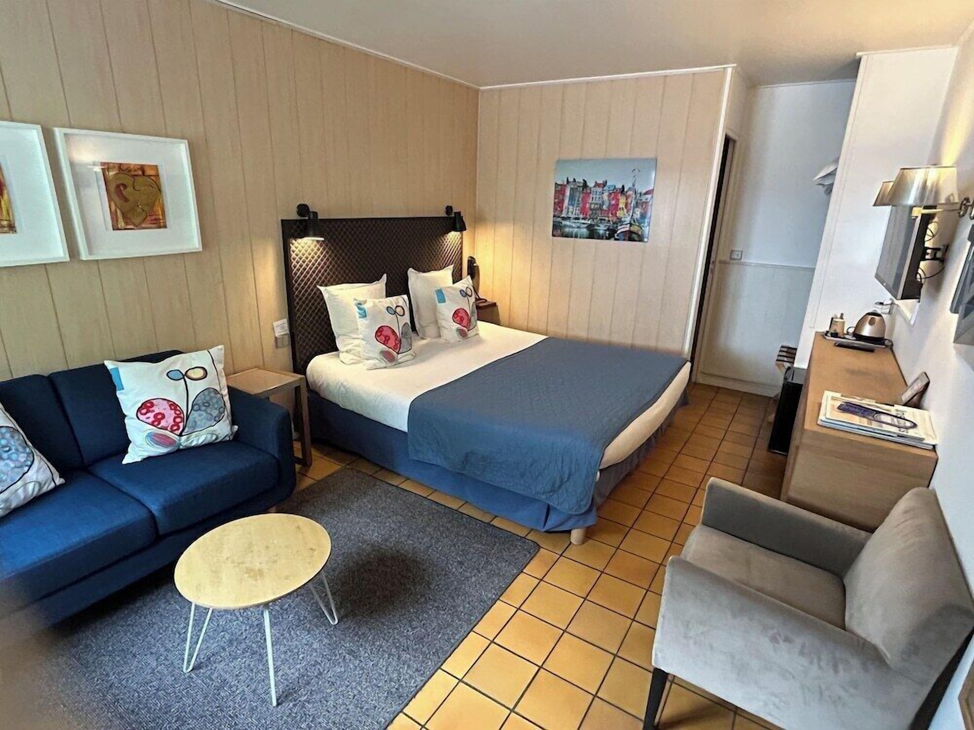 Una habitación de hotel con una cama, un sofá, una silla, una mesita y obras de arte en las paredes.