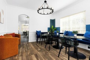 Un espace de salle à manger moderne avec un plancher en bois, des chaises noires et une table ronde. On y trouve des coins salon avec des sièges recouverts de tissu bleu et un lustre au-dessus de la table.