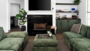 A living room with a fireplace, green sofas, a flat-screen TV, and a kitchen area with shelves. - Luxury 7BR Pool, Hot Tub & 4 Car Parking (Pflugerville)