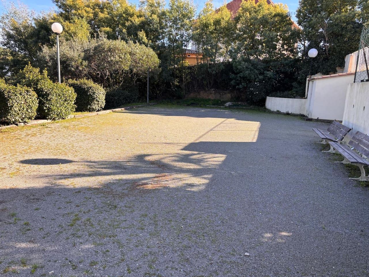 Área externa com piso pavimentado, fileira de bancos e um conjunto de arbustos.