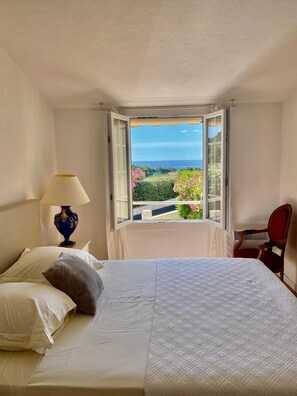 A bedroom with a bed, a nightstand, a lamp, a chair, and a window with a view of the sea and greenery. - Villa Bella – Secure Domaine de la Croisette, Gulf of Saint-Tropez View (Sainte-Maxime)