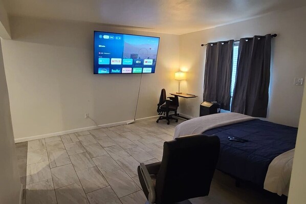 A hotel room with a bed, a desk, a chair, a TV mounted on the wall, and a window with curtains.