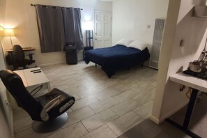 Habitación de hotel pequeña con cama, escritorio con silla, mesita, silla, cortina, puerta y un área de kitchenette.