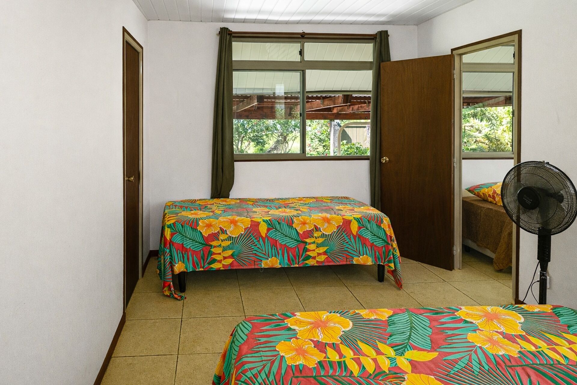 A small room with two beds, a window, and a standing fan.