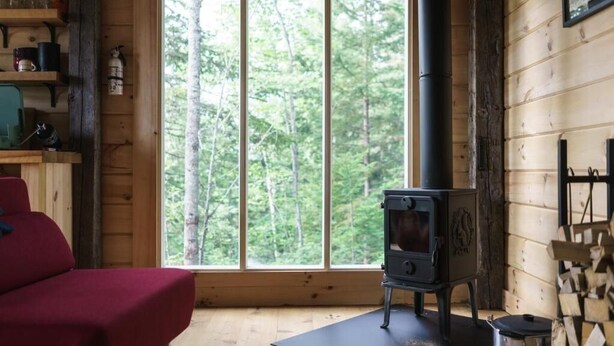 A cozy cabin interior with a large window, a red sofa, a wood-burning stove, and a fireplace mantel. - Les Cabines St-O - 2 (St-Onésime d'Ixworth)