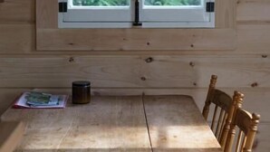 A wooden cabin interior with a table and chairs, a window with a view of greenery, and a small table with a book and a mug. - Les Cabines St-O - 2 (St-Onésime d'Ixworth)
