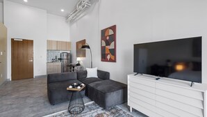 A modern living room with a grey sectional sofa, a white TV stand, and a kitchen in the background. - Studio | near Aquarium, Coolridge Park (Chattanooga)