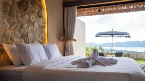 A hotel room with a stone wall, a bed with white linens, a large window with a view of mountains and a body of water, and a beach umbrella outside. - Mount Valley Inn Kintamani (Kintamani)