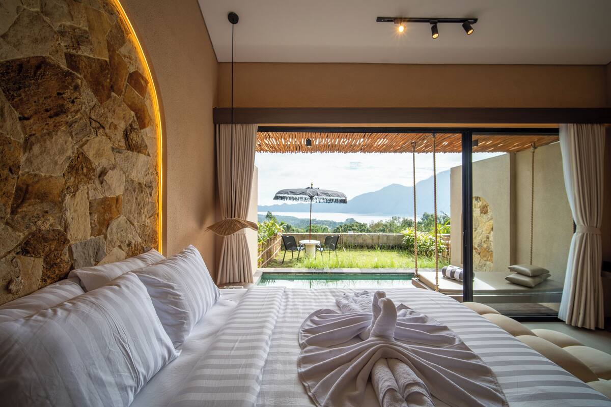Une chambre dotée d’un grand lit, d’un mur en pierre, d’une vue sur une piscine et les montagnes, et d’une porte-fenêtre coulissante.