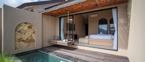 Un área de piscina moderna al aire libre con una terraza de madera, un muro de piedra y un dormitorio con paredes de vidrio, cama y cortinas.