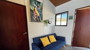A room with a blue sofa, two yellow cushions, a wooden door, a painting of a bird, a window, and a plant. - Quinta Los Monos : Private Swimming Pool : Pet-friendly (Paquera)