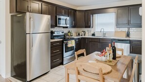Una cocina moderna con muebles oscuros, electrodomésticos de acero inoxidable, una mesa de comedor con sillas y una ventana con persianas.