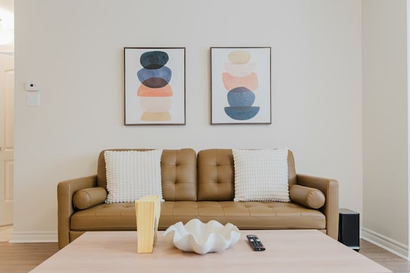 A living room with a brown leather sofa, two framed abstract paintings, a white textured table, a wooden sculpture, a remote control, and a black speaker.