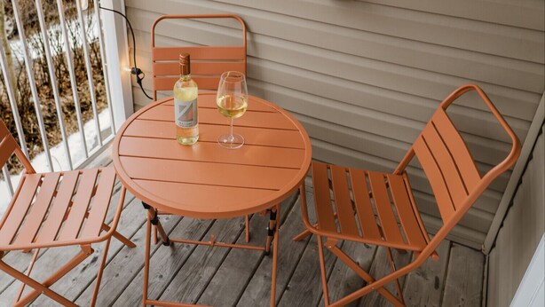 Une table ronde en métal, deux chaises, une bouteille de vin et un verre, le tout posé sur une terrasse en bois.