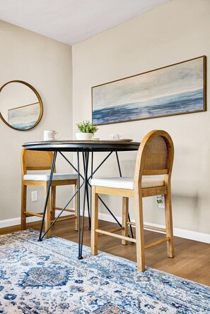 A dining area with a round table, two wooden chairs, a blue patterned rug, a wall-mounted mirror, and a large abstract painting.
