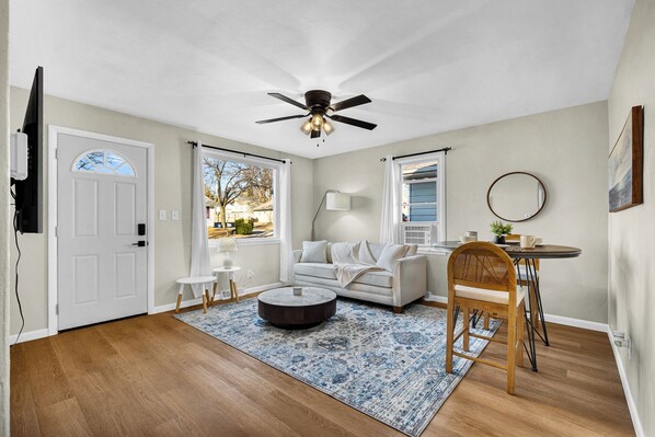 A living room with a ceiling fan, a white couch, a round coffee table, a dining table with chairs, and a large window with curtains.