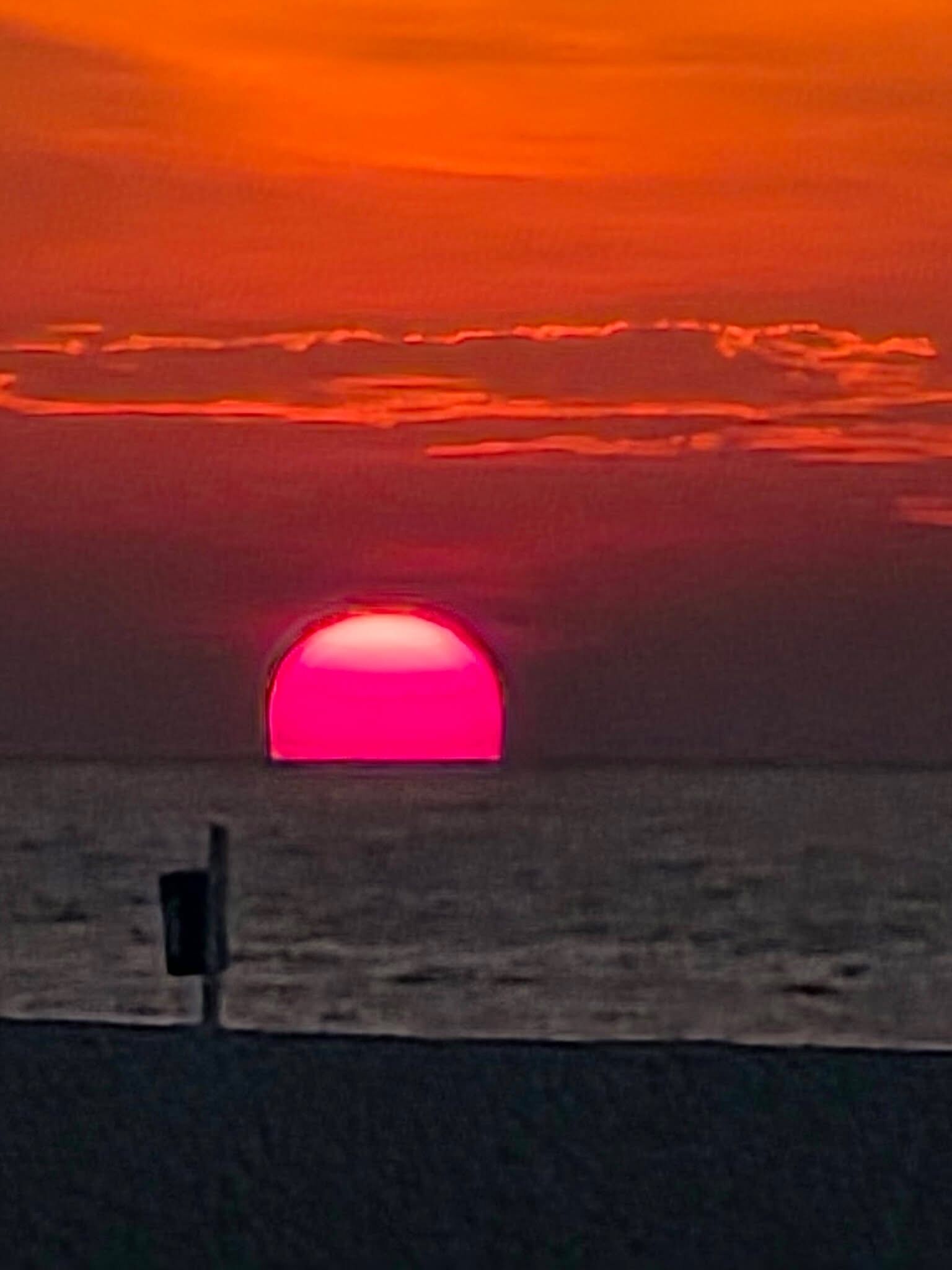 Ein Sonnenuntergang über einem Gewässer, im Vordergrund die Silhouette eines Gebäudes.