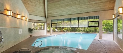 An indoor swimming pool with a hot tub, surrounded by windows and a wooden ceiling.