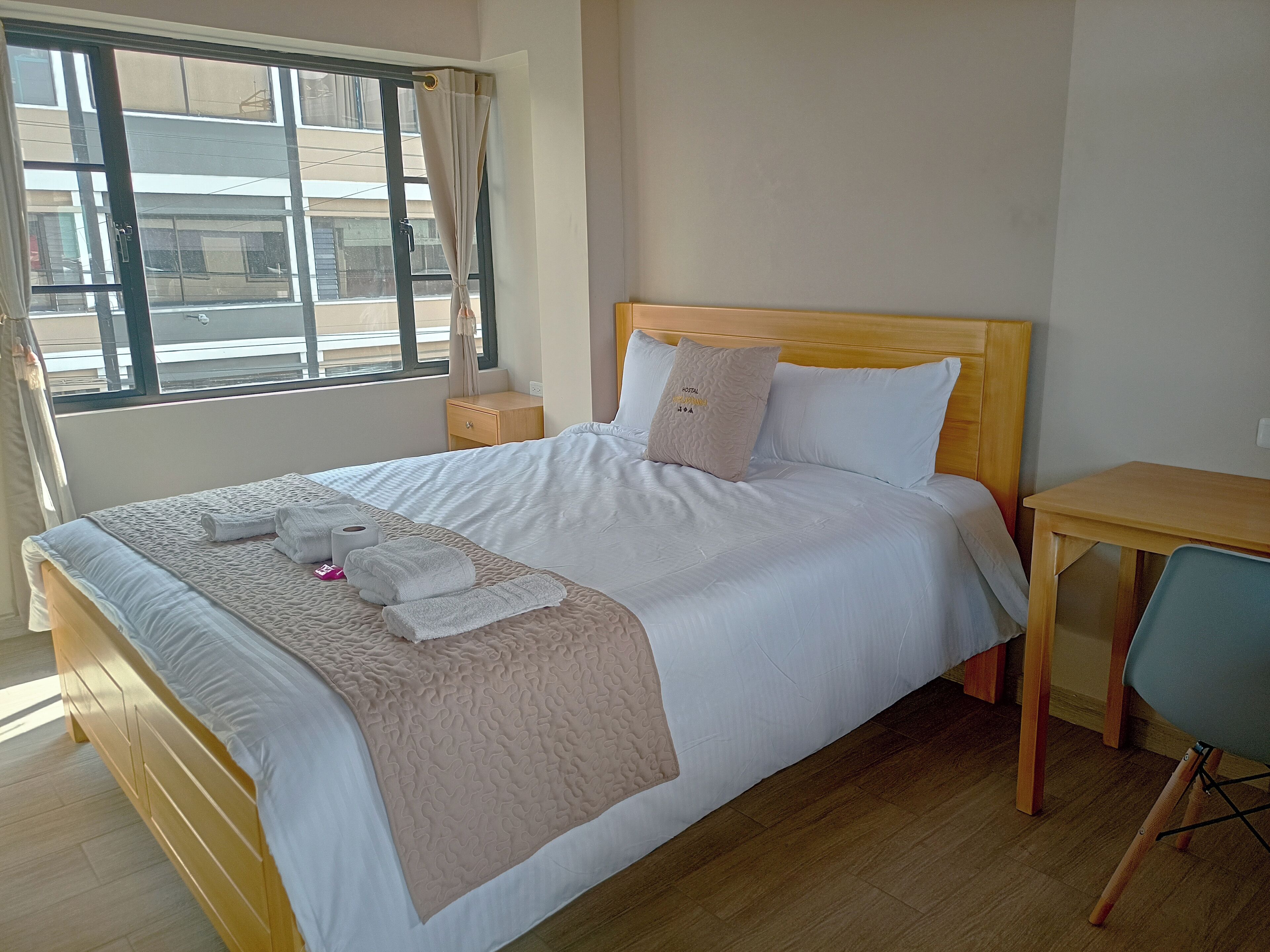 A neatly made bed with a beige quilt, white pillows, and a "HOSTAL" pillow. There is a wooden nightstand with a lamp, a wooden desk, and a chair. The room has a large window with curtains, wooden flooring, and a view of buildings outside.