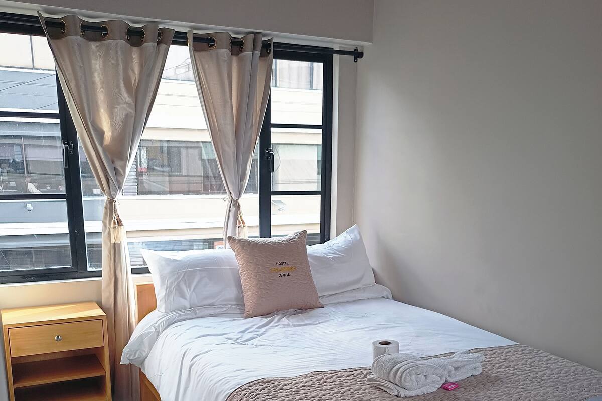 Een net opgemaakt bed met een beige deken, een houten nachtkastje met lade en een raam met gordijnen.