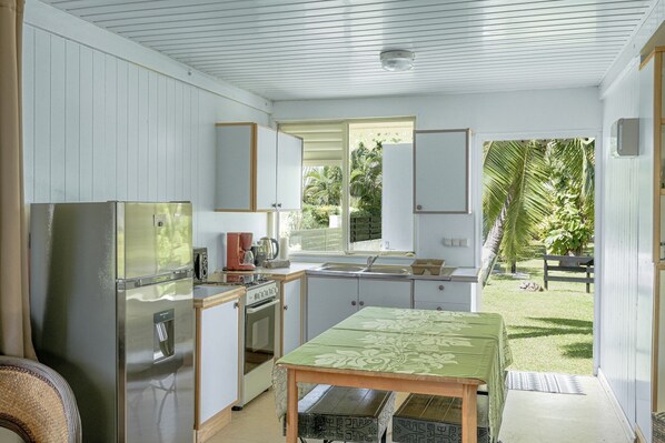 A compact kitchen with a refrigerator, stove, and sink. There is a table with a green tablecloth and a view of a garden outside.