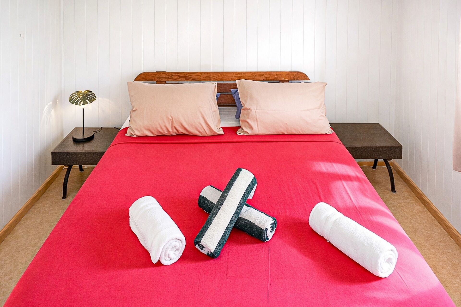 A neatly made bed with a red bedspread, two pillows, and three rolled towels.