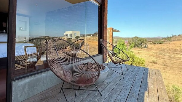 Une terrasse en bois avec deux fauteuils en osier, une porte vitrée donnant sur une pièce avec un lit et une table, et une vue sur un paysage désertique.