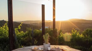 Une terrasse en bois avec une table en pierre et quatre tabourets, offrant une vue panoramique sur un paysage vallonné et un coucher de soleil.