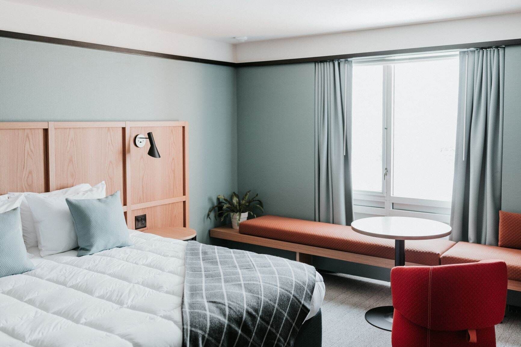 A hotel room with a bed, a bench, a red chair, a small table, and a window with curtains.