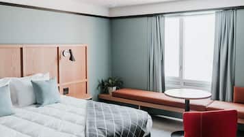 A hotel room with a bed, a bench, a red chair, a small table, and a window with curtains.