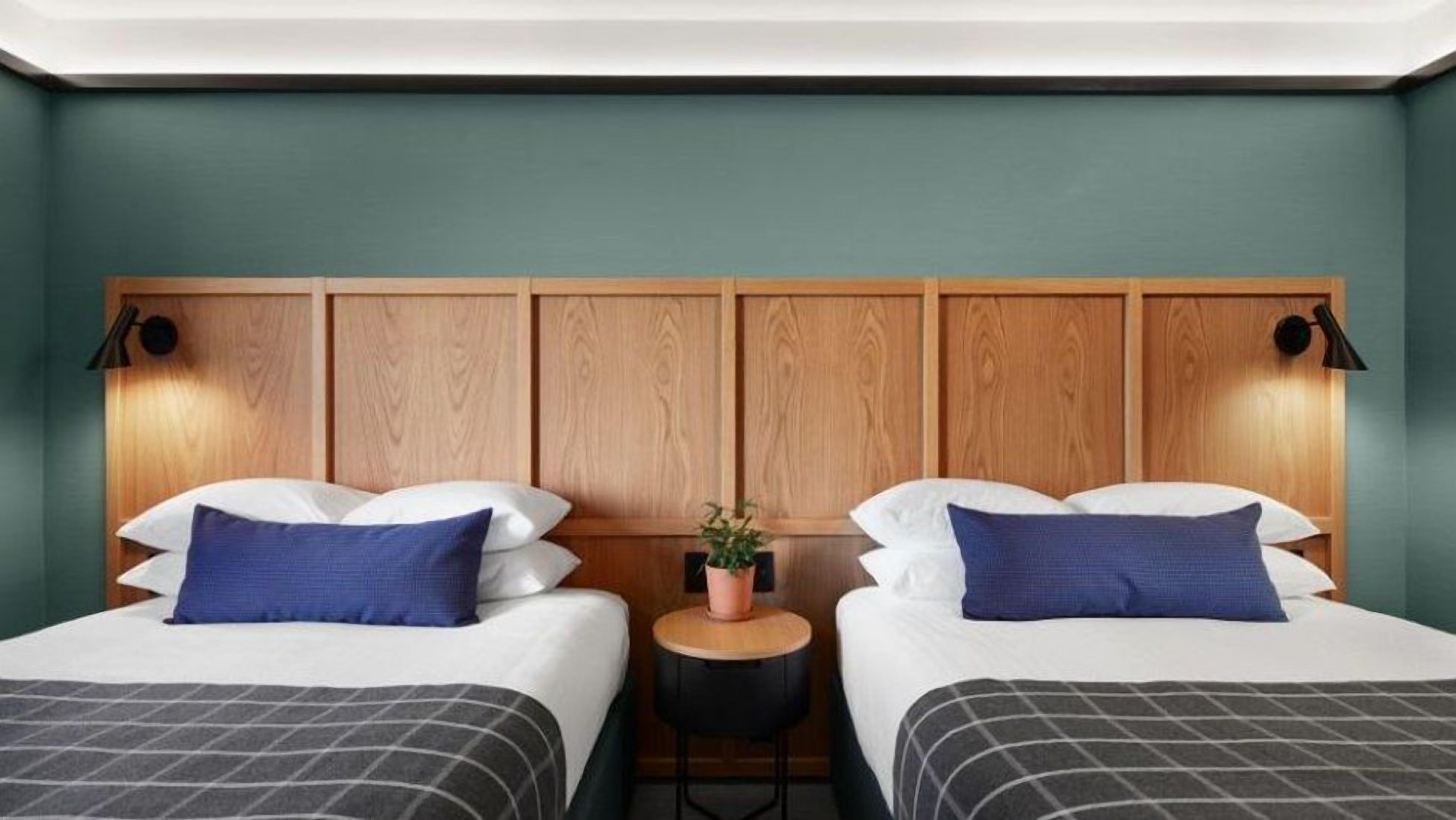 A hotel room with two beds, a wooden headboard, bedside lamps, a small round table with a plant, and a potted plant on the floor.