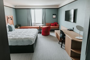 A hotel room with a bed, a desk, a chair, a sofa, a TV, and a window with curtains. - Haworth Inn & Conference Center (Holland)