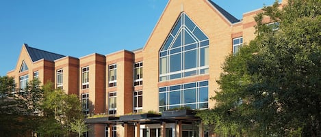 A modern building with a prominent triangular glass facade, a covered entrance area with seating, and a well-maintained surrounding landscape.