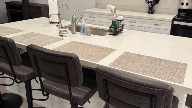 Una cucina moderna con isola centrale, sgabelli da bar, mobili bianchi, lavello in acciaio inossidabile e televisione a schermo piatto montata a parete.
