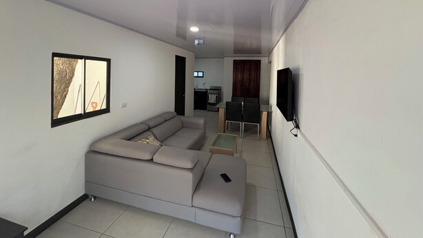 A modern living room with a sectional sofa, a dining area with a table and chairs, a flat-screen TV mounted on the wall, and a kitchen in the background. - Floral Departments (Comunidad)