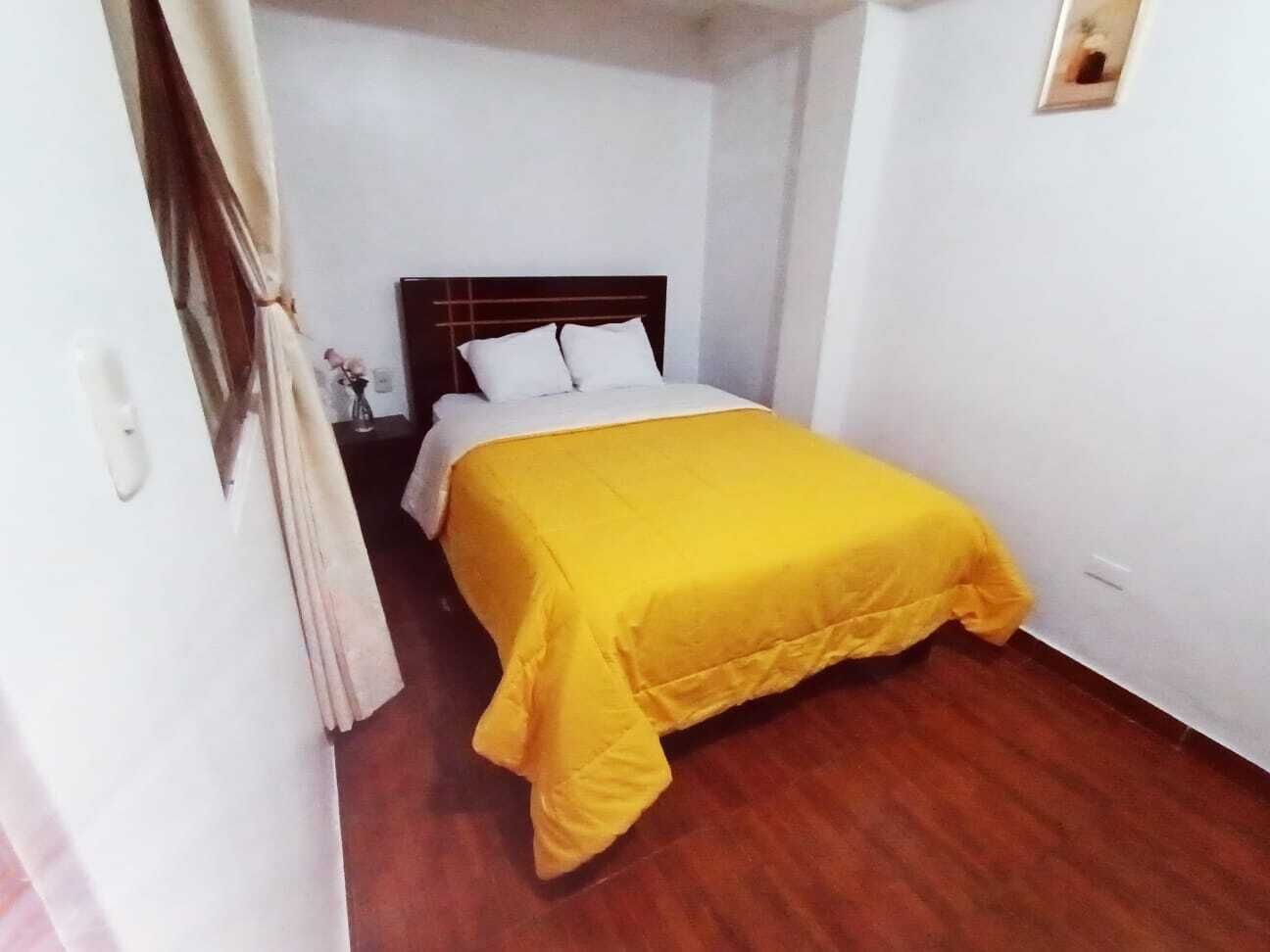 Un dormitorio con piso de madera, cama con cubrecama amarillo, almohadas blancas y cabecera de madera.