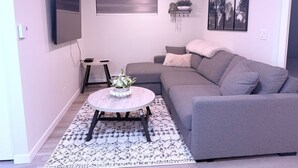 Una sala de estar moderna con un sofá seccional gris, una mesa redonda de centro con un arreglo floral, un televisor de pantalla plana montado y una obra de arte con una vista urbana enmarcada en la pared.