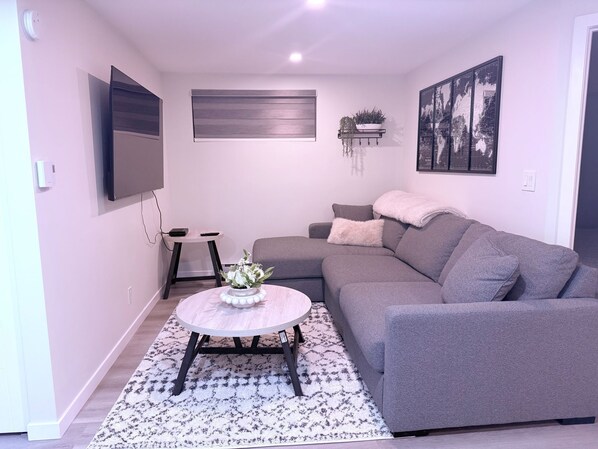 A modern living room with a grey sectional sofa, a round coffee table with a floral centerpiece, a mounted flat-screen TV, and a framed cityscape artwork on the wall.