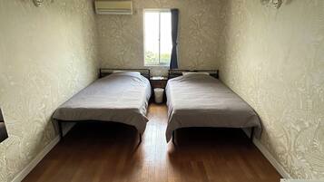 A room with two beds, a window with curtains, an air conditioning unit, and a wooden floor.