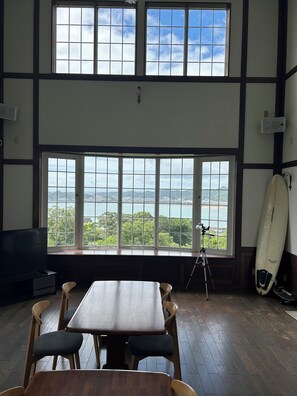 Una habitación con un ventanal grande que da a una masa de agua, una mesa de comedor con sillas y una tabla de surf apoyada contra la pared.