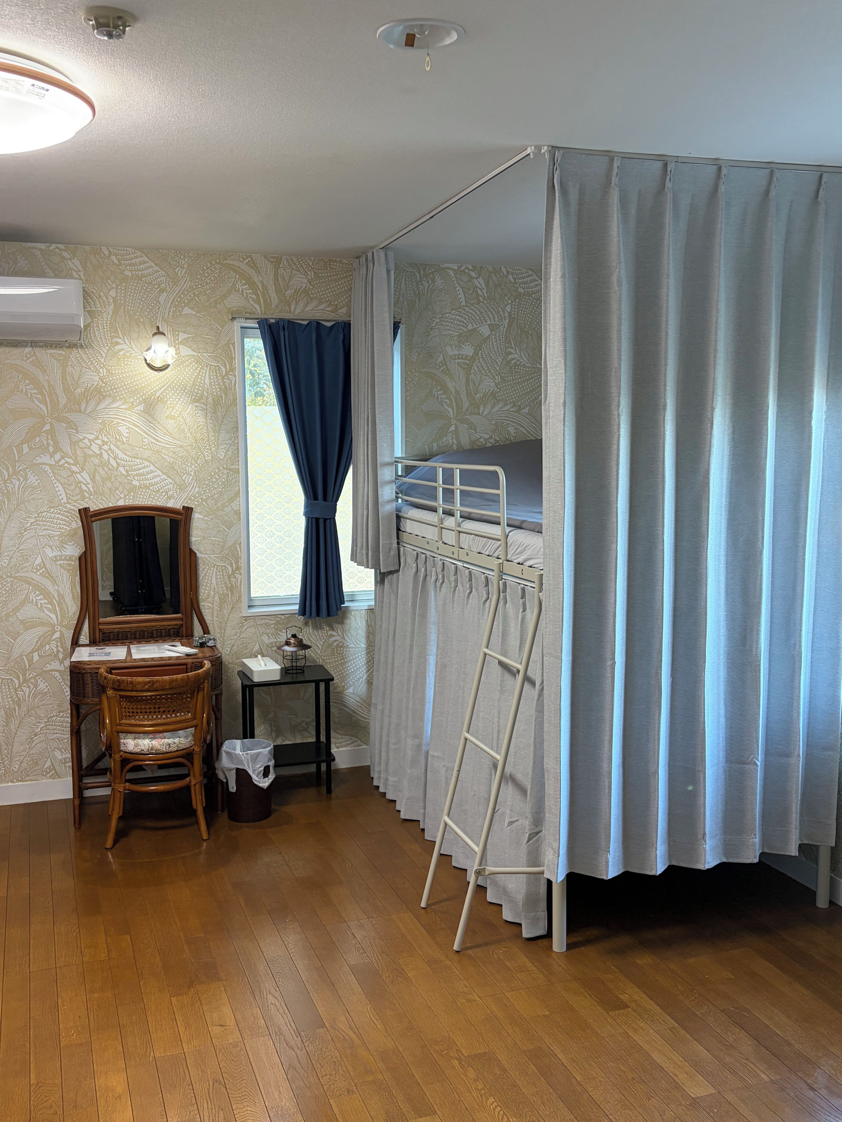 Een kamer met een stapelbed, een stoel, een klein tafeltje, een ladder en een raam met gordijnen.