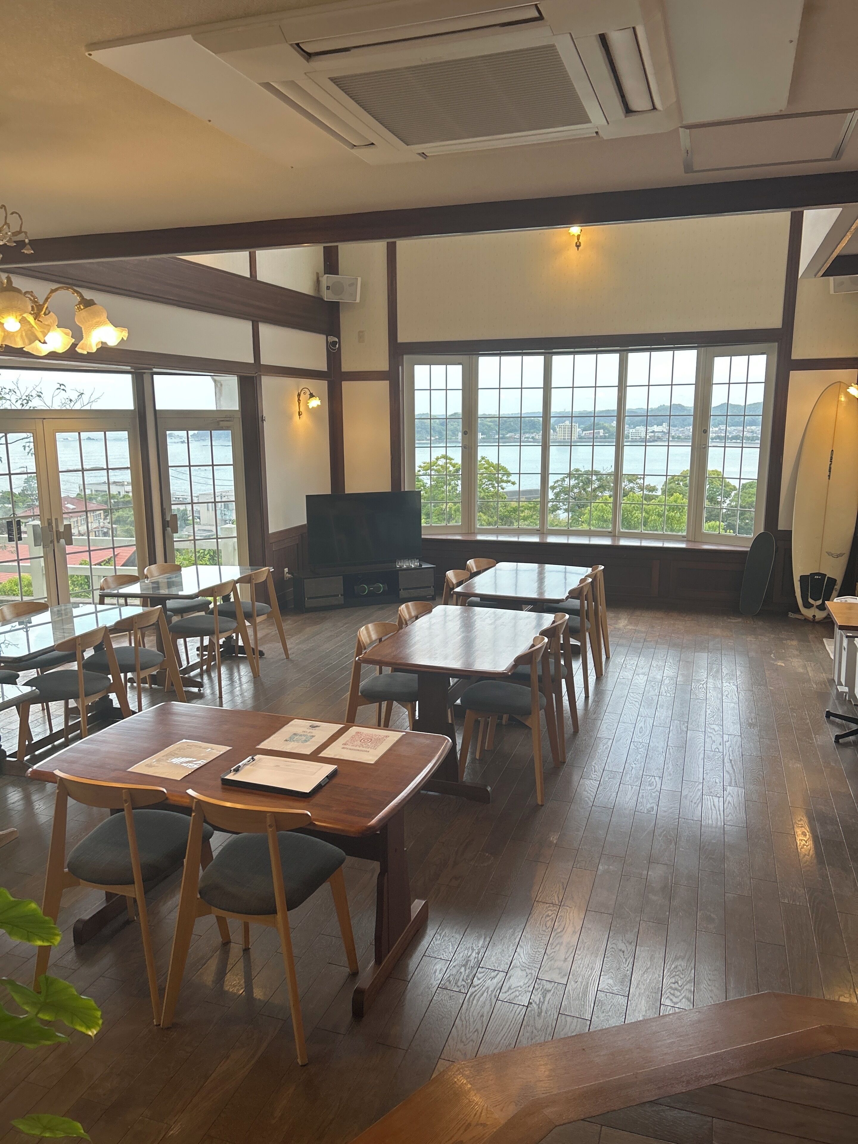 A restaurant with wooden tables and chairs, large windows with a view of the sea, and a surfboard on the wall.