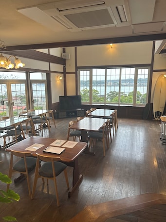 A restaurant with wooden tables and chairs, large windows with a view of the sea, and a surfboard on the wall.