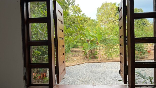 Une porte en bois ouverte donne sur un chemin de gravier bordé d’une végétation luxuriante et de bananiers.