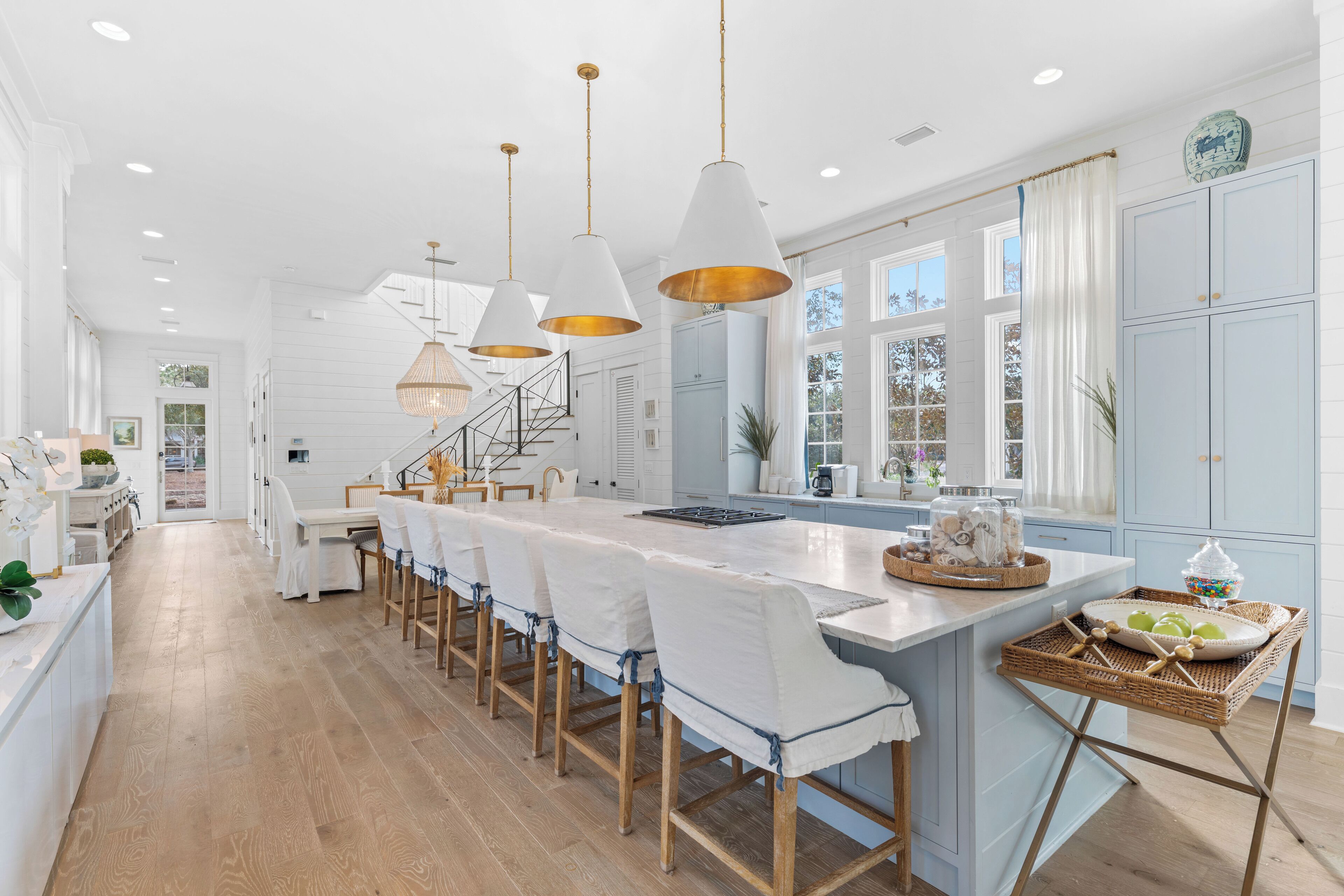 A spacious kitchen with a long island, white chairs, pendant lights, and a view of the outdoors.