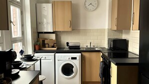 A compact kitchen with wooden cabinets, a washing machine, a microwave, and a sink. - Heart of London Signature Stay | Sleeps 6 (Greater London)