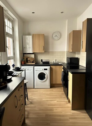 A compact kitchen with wooden cabinets, a washing machine, a microwave, and a sink. - Heart of London Signature Stay | Sleeps 6 (Greater London)